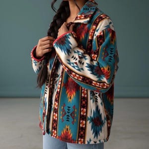 Op de afbeelding: Een fleece jack met een patroon geïnspireerd op het zuidwesten. De jas heeft een geometrisch ontwerp in teal, rood, oranje en crème. De kraag en manchetten hebben een rode bies. De jas wordt gedragen over een blauwe spijkerbroek.