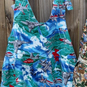 May include: A blue and green sleeveless dress with a white background. The dress is covered in a pattern of fighter jets. The dress has a ruffled hem and two straps that tie at the shoulders.