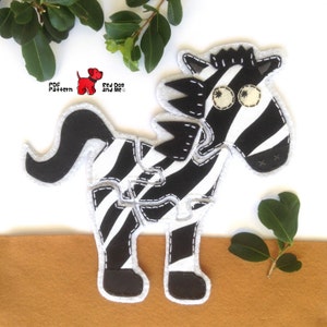 Puede incluir: Juguete de cebra de fieltro negro y blanco con una cara y ojos cosidos. El juguete está hecho de varias piezas que se cosen juntas. El juguete está sobre un fondo de fieltro marrón. El texto "PDF Pattern Red Dog and Me" está en la esquina superior izquierda de la imagen.