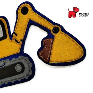 Può includere: Un escavatore in feltro giallo, blu, grigio e marrone. L'escavatore ha una cabina, un braccio e una benna. Il design include dettagli cuciti e un logo di un cane rosso con la scritta "Red Dog and Me".
