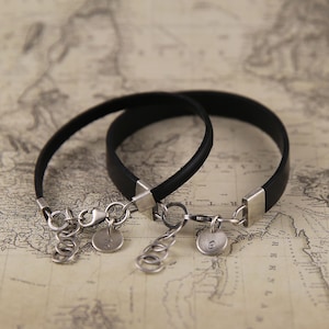May include: Two black leather bracelets with silver clasps and round silver charms. The charms have the numbers "24" and "63" engraved on them.