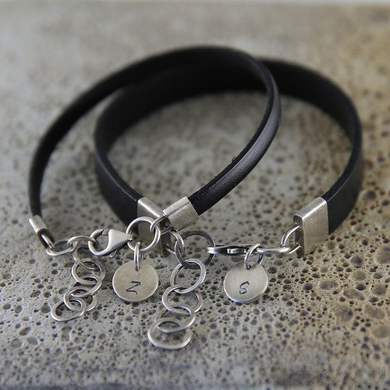 May include: Two black leather bracelets with silver clasps and charms. The charms are round and have the letters "N" and "S" engraved on them.