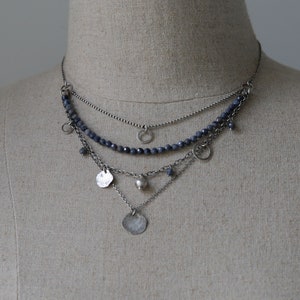 May include: A layered silver necklace with a beaded chain and multiple charms, including small circles, a round bead, and two flat, textured discs.
