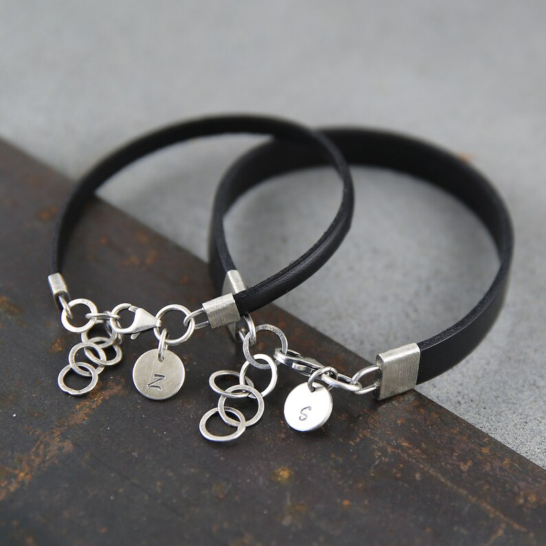 May include: Two black leather bracelets with silver clasps and charms. The charms are round and have the letters "N" and "S" engraved on them.