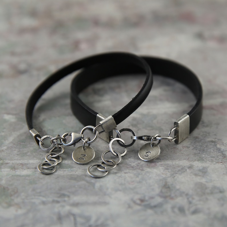 May include: Two black leather bracelets with silver clasps and charms. The charms are round and have the letters "N" and "S" engraved on them.