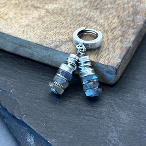Puede incluir: Pendientes de aro plateados con cuentas de piedras preciosas azules y grises facetadas y apiladas colgando. Los pendientes se muestran sobre una superficie de madera con un fondo gris oscuro.