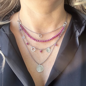 May include: A multi-strand necklace with a silver finish. The necklace features a strand of small, faceted, magenta beads, along with dangling silver circles and hammered silver coin pendants. The necklace is worn against a black shirt.