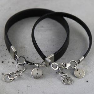 May include: Two black leather bracelets with silver clasps and charms. The charms are round and have the letters "S" and "N" engraved on them.