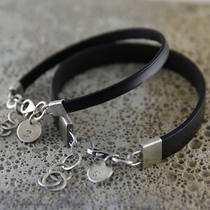 May include: Two black leather bracelets with silver clasps and charms. The charms are round and have the letters "N" and "S" engraved on them.