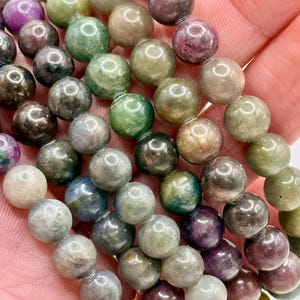 May include: Strands of gemstone beads in various colors, including green, purple, and gray. The beads are round and appear to be polished. The image shows a close-up view of the beads, highlighting their texture and color variations. These beads are suitable for jewelry making.