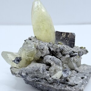 以下が含まれることがあります： 大きな淡い黄色の結晶が特徴の鉱物標本。結晶は、より小さく透明で灰色の結晶に囲まれ、灰色のテクスチャードマトリックス上にあります。暗く金属的な外観の結晶も存在します。