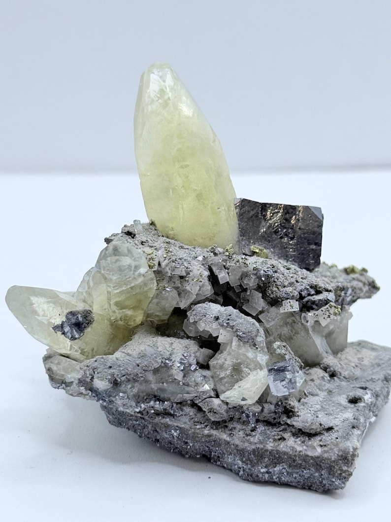 以下が含まれることがあります： 大きな淡い黄色の結晶が特徴の鉱物標本。結晶は、より小さく透明で灰色の結晶に囲まれ、灰色のテクスチャードマトリックス上にあります。暗く金属的な外観の結晶も存在します。
