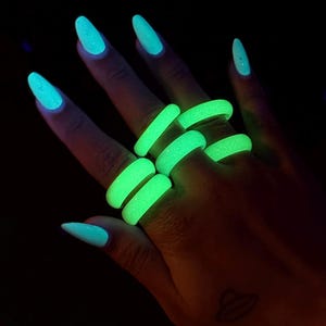 May include: A hand wearing several glowing green rings. The rings are a bright, neon green color and appear to be made of a rubbery material. The nails are painted a light blue color. The background is black.
