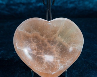 May include: A heart-shaped pink Himalayan salt dish on a black metal stand. The dish is made of natural salt crystals and has a rough, textured surface.