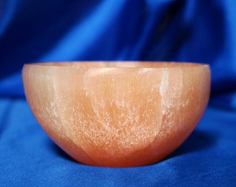 May include: A peach-coloured, semi-translucent bowl with a rounded shape. The bowl has a smooth exterior and a slightly textured interior. The background is a solid blue fabric.