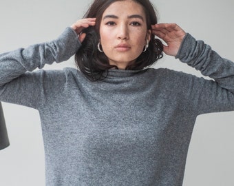 May include: A woman wearing a gray long-sleeved sweater with a mock neck and black pants.
