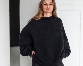 May include: A woman wearing a black oversized sweater and black wide-leg pants. She is standing in a white room with white pillars.
