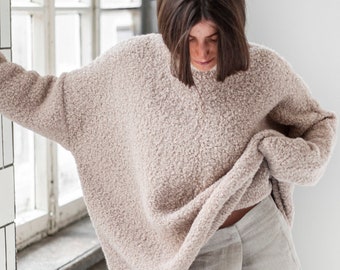 May include: A woman wearing a light brown fuzzy sweater and light gray linen pants. She is standing in a room with white tiled walls and a window.