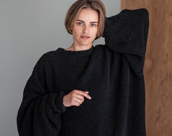 May include: A woman wearing a black fuzzy sweater with long sleeves and a relaxed fit. She is standing in front of a wooden wall.