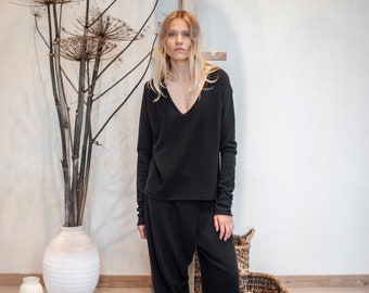 May include: A woman wearing a black long-sleeved v-neck sweater and black pants with a wide leg. She is wearing black clogs and standing in front of a white wall.
