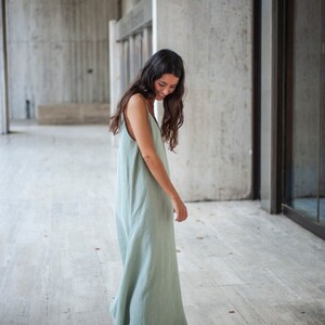 May include: A woman wearing a sage green linen maxi dress with a simple, sleeveless design. The dress has a flowing silhouette and is slightly gathered at the waist. She is standing on a light-colored tiled floor.