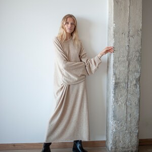 May include: A woman wearing a long, beige sweater dress with long sleeves and a relaxed fit. She is standing next to a concrete pillar and wearing black ankle boots.