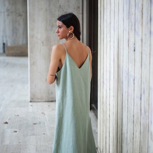 May include: A woman wearing a sage green maxi dress with thin straps and a V-neckline. She is standing in front of a concrete wall.