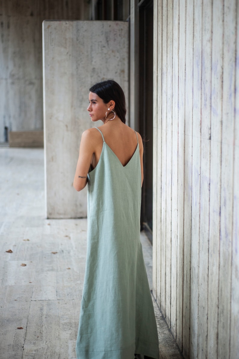 May include: A woman wearing a sage green maxi dress with thin straps and a V-neckline. She is standing in front of a concrete wall.
