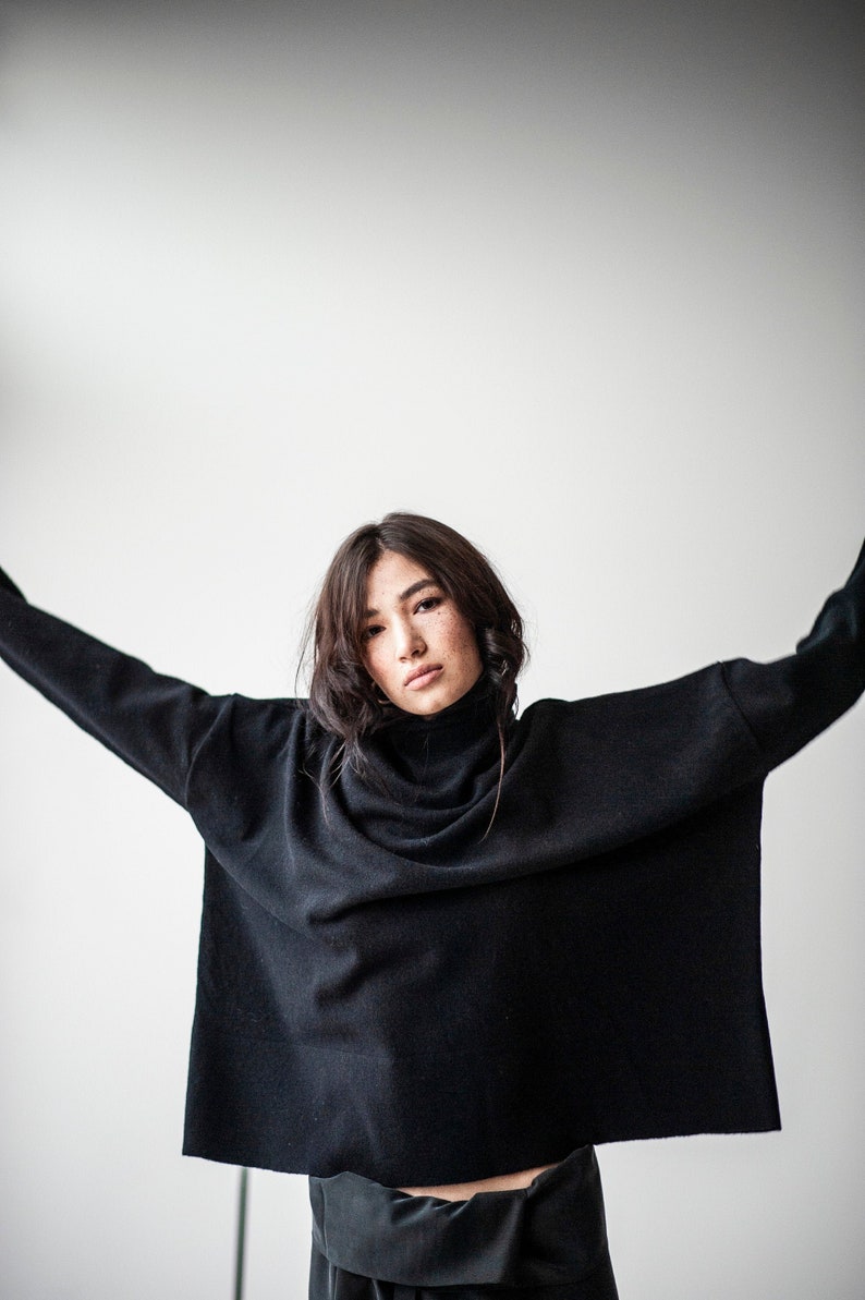 May include: A woman wearing a black turtleneck sweater with her arms outstretched. The sweater is loose fitting and has a relaxed, casual style.