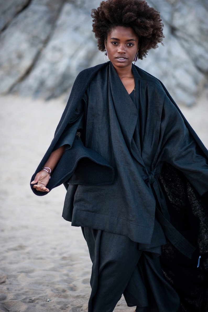 May include: A woman wearing a black, long-sleeved, wrap-style top with a flowing silhouette. The top has a wide, open collar and a belt at the waist. The woman has dark curly hair and is standing on a sandy beach.