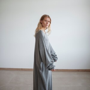 May include: A woman wearing a long, gray, oversized sweater dress. She is standing in a white room with a concrete floor.