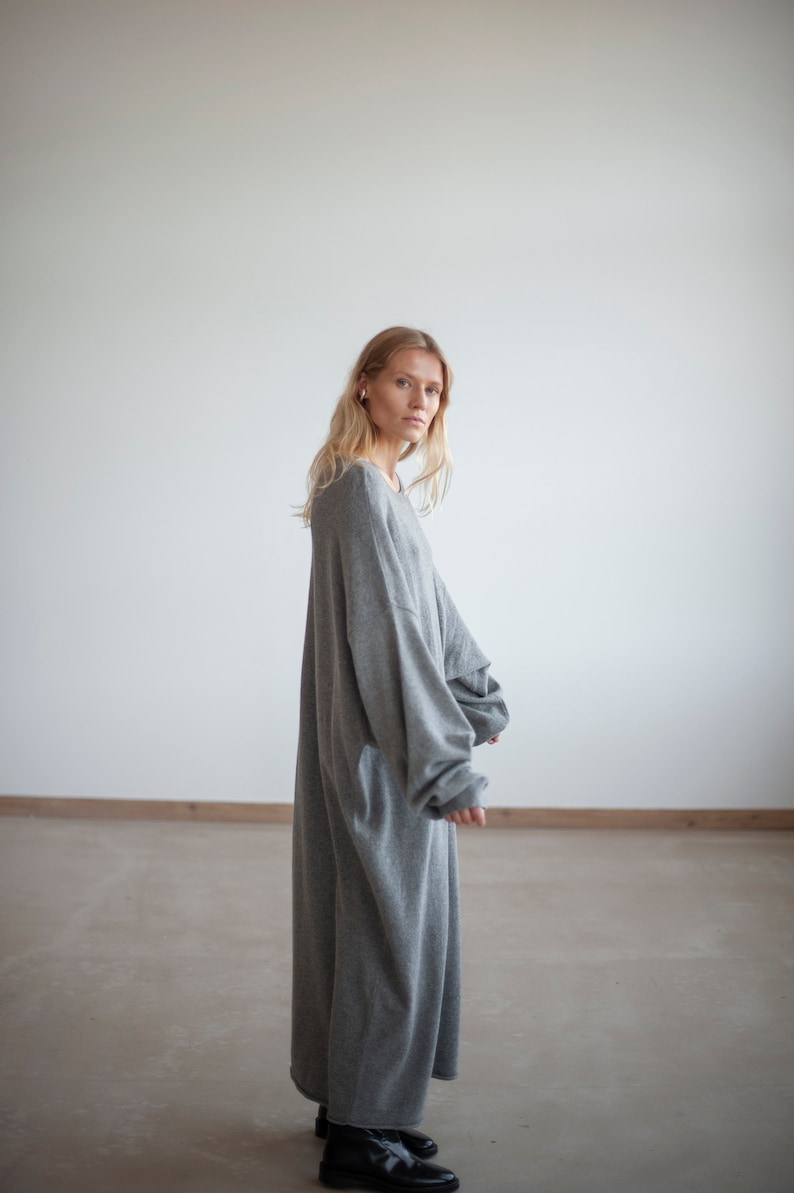 May include: A woman wearing a long, gray, oversized sweater dress. She is standing in a white room with a concrete floor.