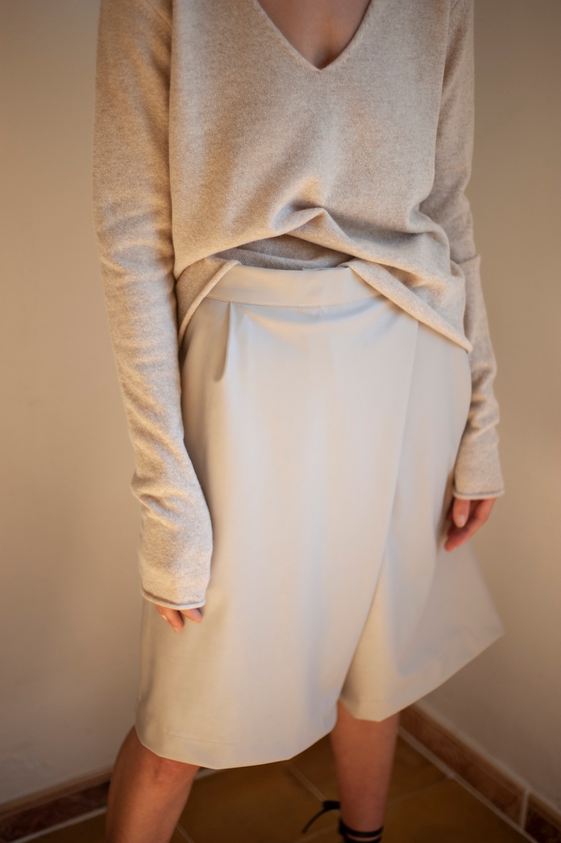 May include: A light beige sweater with long sleeves and a v-neck. The sweater is worn over a light gray skirt with a wrap-style design.