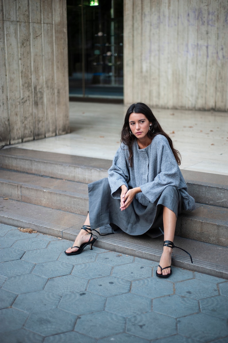 May include: A woman wearing a grey and white patterned sweater and a grey skirt sits on a set of concrete steps. She is wearing black sandals with straps.