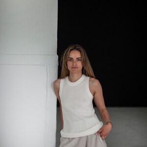 Op de afbeelding: Een vrouw in een wit geribbeld tanktop en een beige wijde broek staat tegen een witte muur met een zwarte achtergrond.