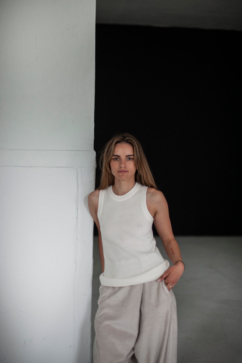 Op de afbeelding: Een vrouw in een wit geribbeld tanktop en een beige wijde broek staat tegen een witte muur met een zwarte achtergrond.