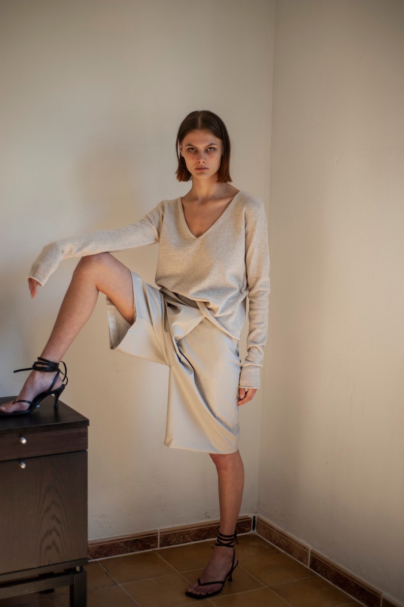 May include: A woman wearing a light beige sweater and matching beige shorts. She is standing with one leg raised and wearing black heeled sandals.