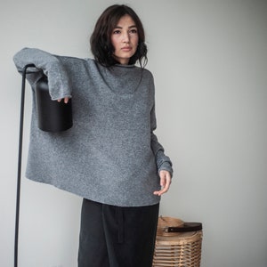 May include: A woman wearing a gray oversized sweater and black pants. She is standing in front of a white wall with a black lamp and a wicker basket.
