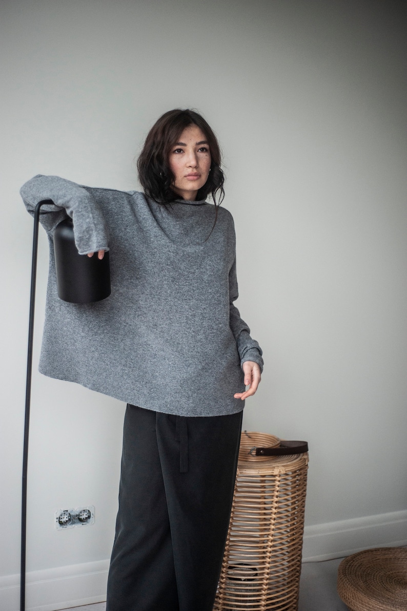May include: A woman wearing a gray oversized sweater and black pants. She is standing in front of a white wall with a black lamp and a wicker basket.