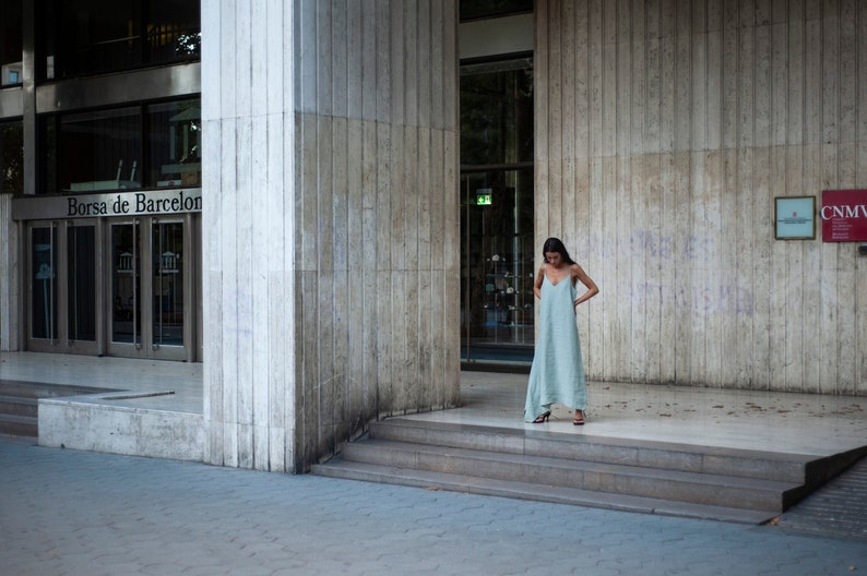 May include: A woman in a light blue maxi dress stands in front of a building with the text "Borsa de Barcelona" on the side. The building has a concrete facade and a large glass door.