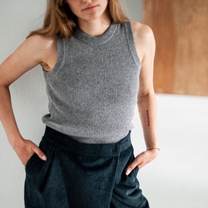 Op de afbeelding: Een vrouw draagt een grijs geribbeld tanktop en een donkergroene corduroy broek. Ze heeft een tatoeage op haar arm die "In Love We Rise" zegt.
