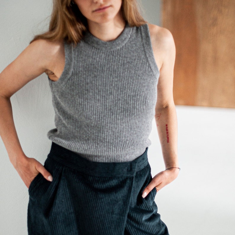 Op de afbeelding: Een vrouw draagt een grijs geribbeld tanktop en een donkergroene corduroy broek. Ze heeft een tatoeage op haar arm die "In Love We Rise" zegt.