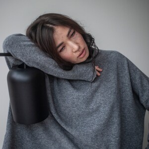 May include: A woman wearing a gray oversized sweater with a relaxed fit. She is leaning against a black lamp with a cylindrical shade.