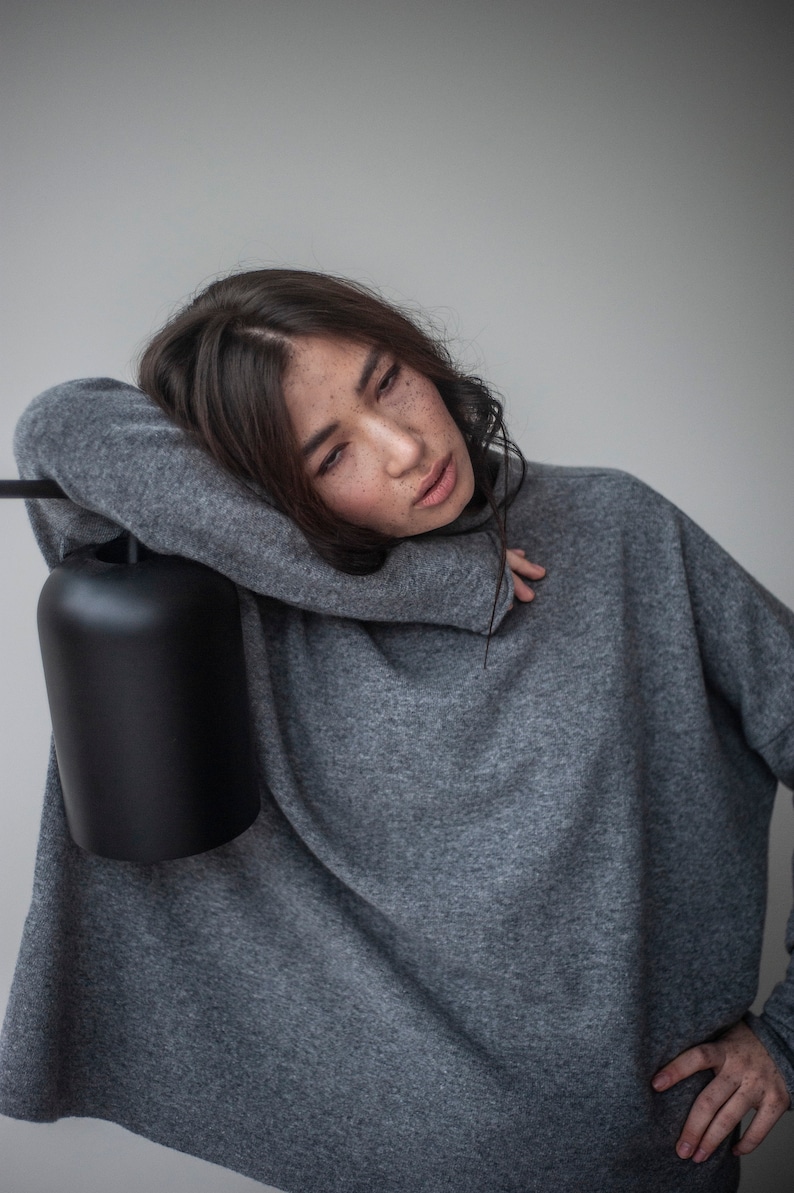 May include: A woman wearing a gray oversized sweater with a relaxed fit. She is leaning against a black lamp with a cylindrical shade.