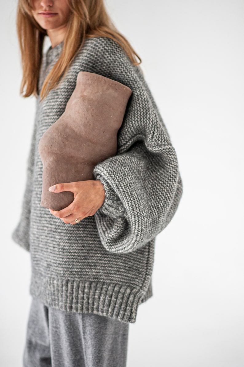 May include: A woman wearing a gray knitted sweater with a brown ceramic vase.