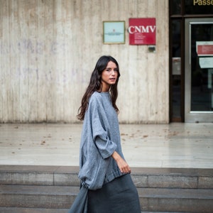 May include: A woman wearing a grey and white patterned sweater and a long grey skirt. She is walking down a set of stairs in front of a building with a sign that reads "CNMV".