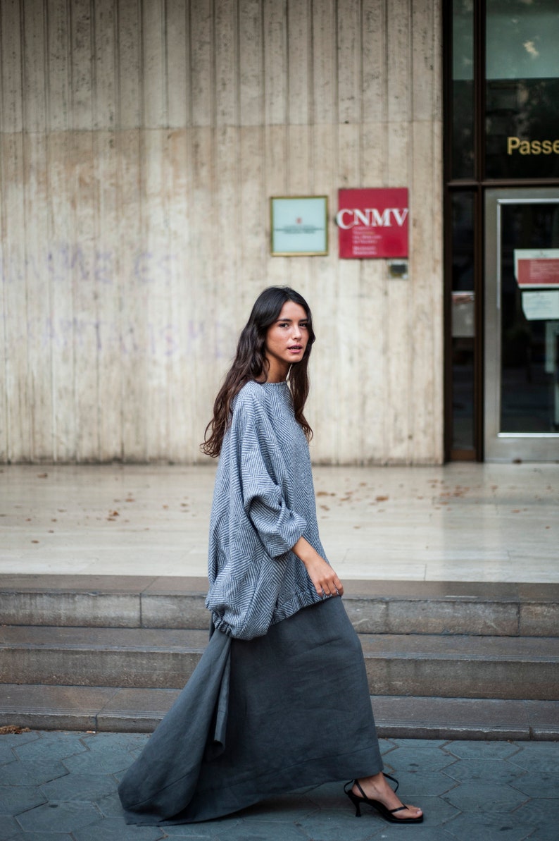 May include: A woman wearing a grey and white patterned sweater and a long grey skirt. She is walking down a set of stairs in front of a building with a sign that reads "CNMV".
