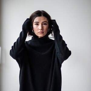 May include: A woman wearing a black mock neck sweater with her hands tucked inside the sleeves.