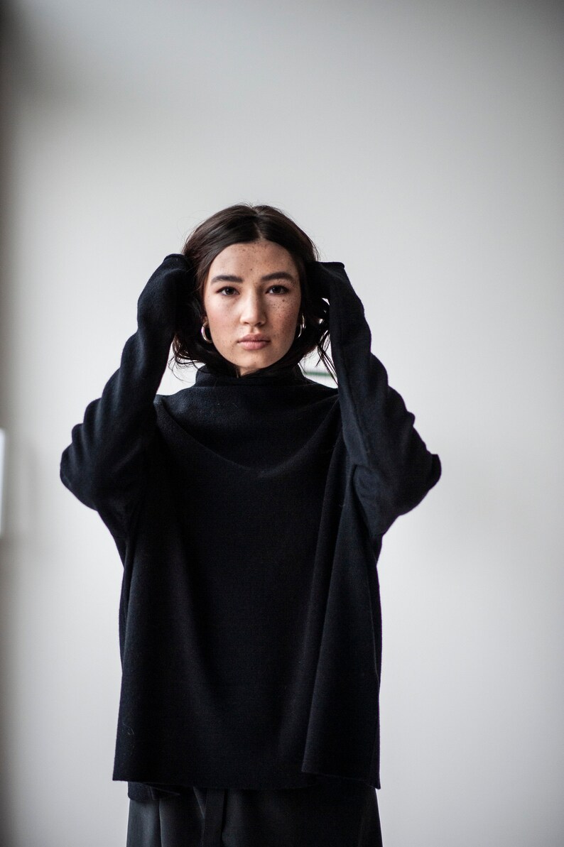 May include: A woman wearing a black mock neck sweater with her hands tucked inside the sleeves.