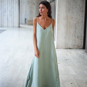 May include: A woman wearing a light green maxi dress with a V-neckline and thin straps. The dress has a flowing, asymmetrical hemline. She is standing in a concrete hallway.